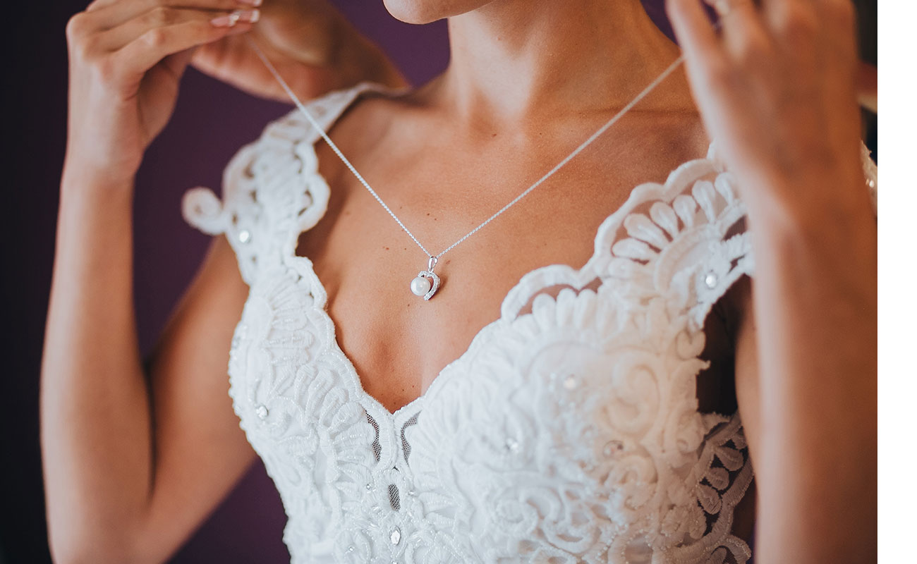 Woman's torso in wedding dress putting on necklace with help from someone behind her.